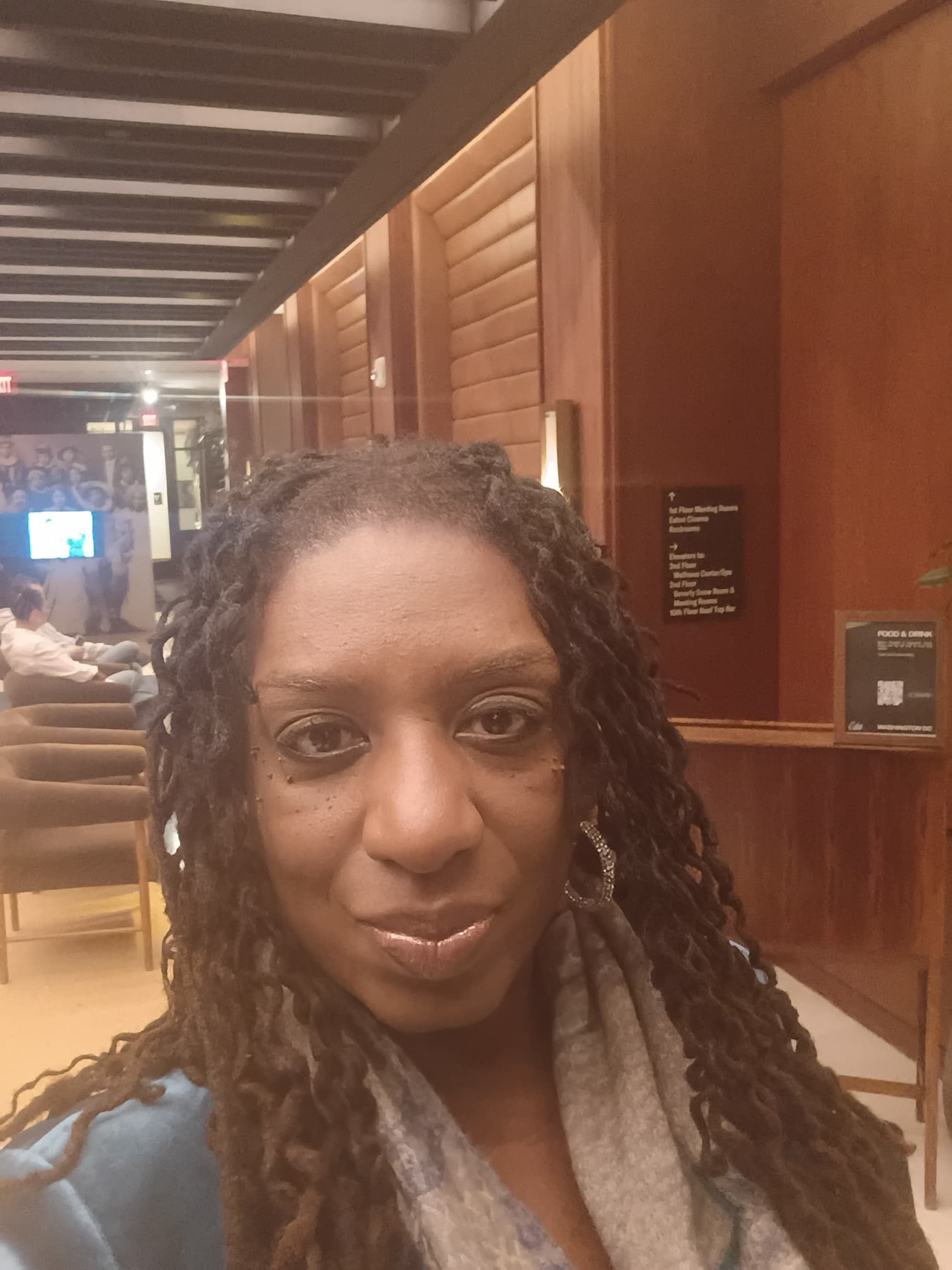Smiling woman with long dark dreadlocks and silver earrings in a wood-paneled indoor setting.
