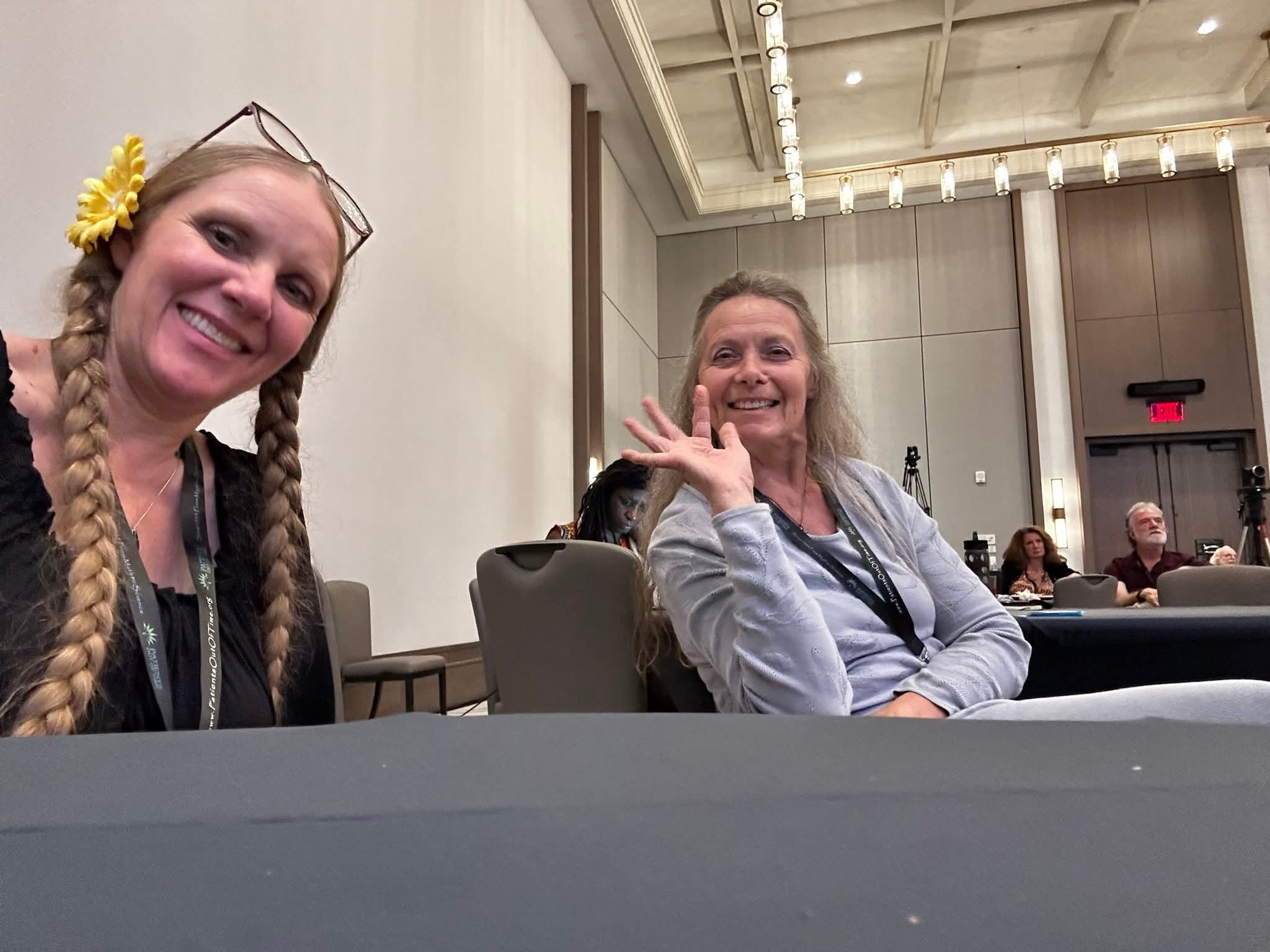 Two smiling women at a conference; one has blonde braids and the other is waving.